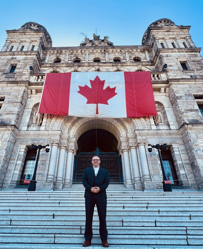 Image Description: Steve standing on the stone steps at the entrance to the B.C. legislature building with a large Canada flag hung above him. He is wearing a dark suit and smiling at the camera.
